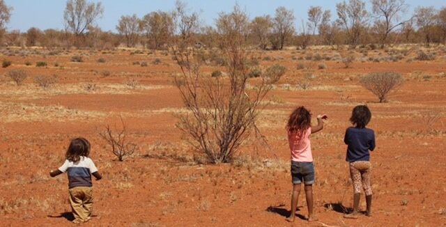 Three young children standing in the red dirt on Martu Country.