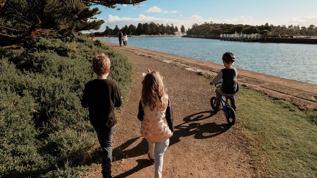 Two kids walking along a lake track and 1 riding a bike, backs all turned to the camera.