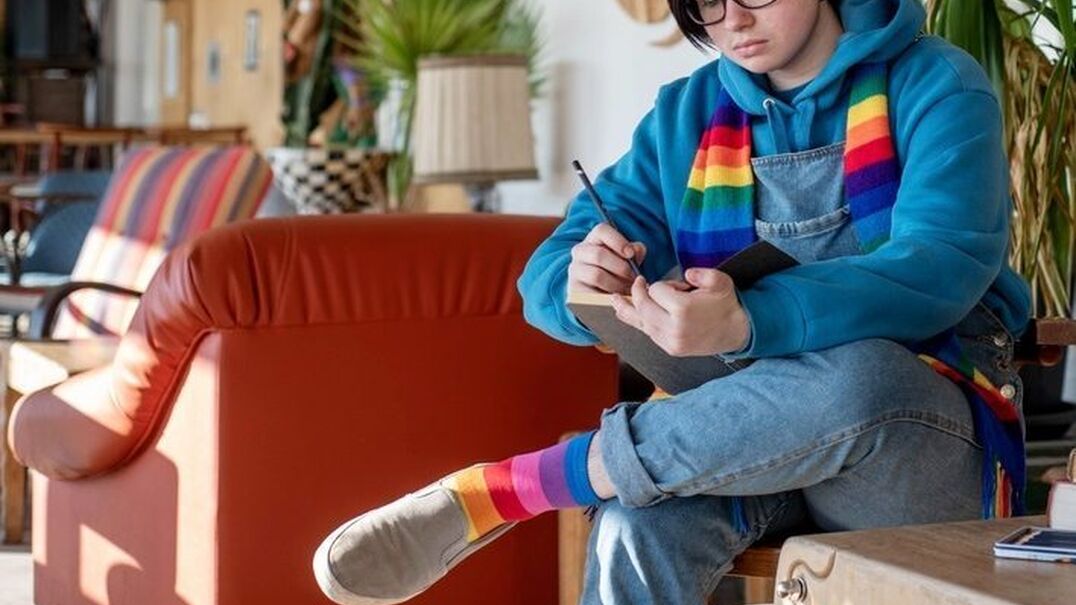 Non binary teen focussed on drawing in an art journal at home and wearing a rainbow scarf and socks.
